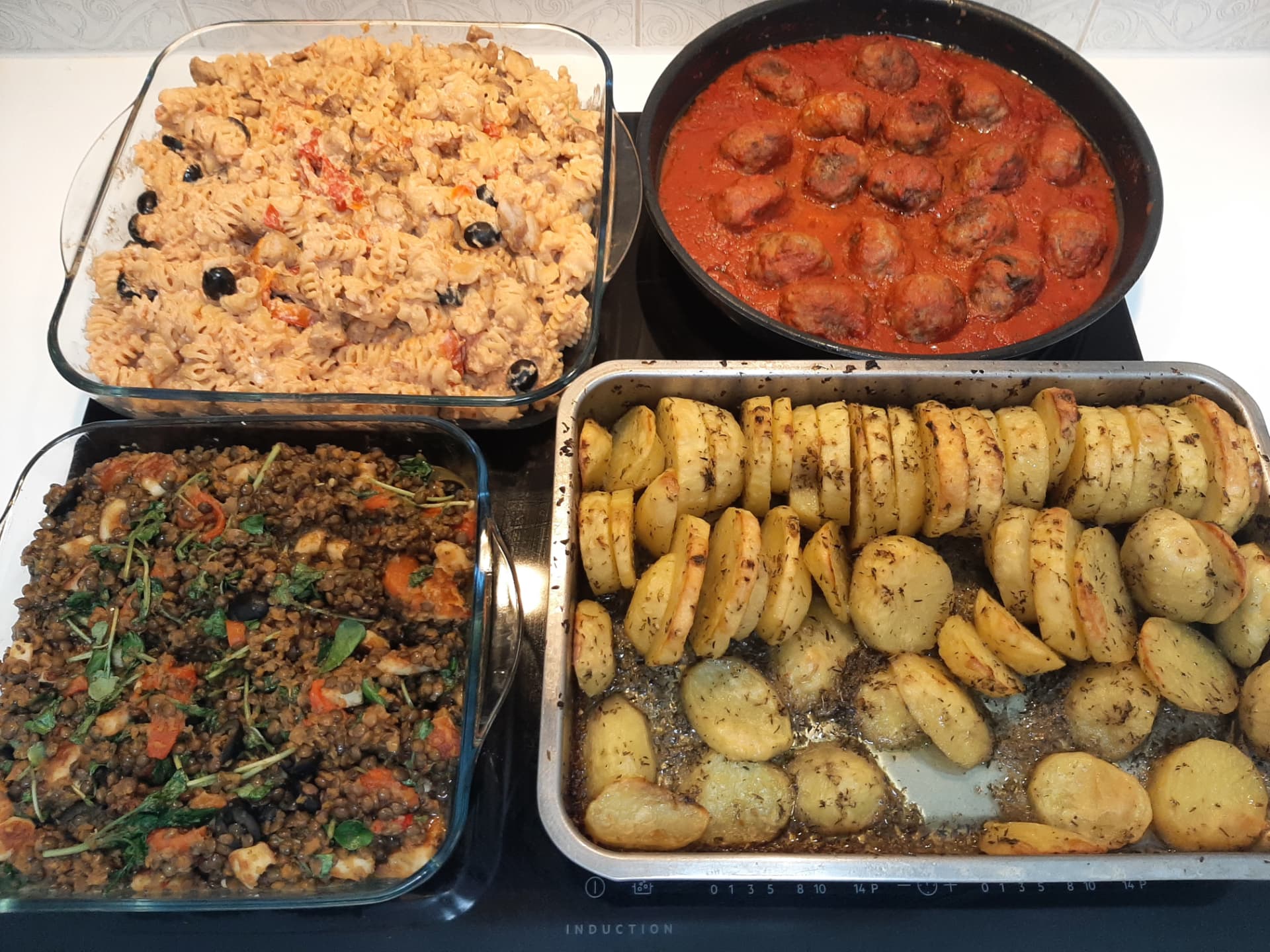 Four freshly cooked dishes ready on the hob: creamy pasta with olives and tomatoes, meatballs in tomato sauce, a lentil salad with vegetables, and a tray of herbed roast potatoes